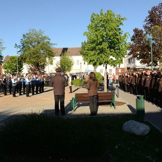 Eine Band tritt im Freien auf, während eine Menschenmenge zusieht. Die Bandmitglieder tragen blaue und weiße Uniformen. Eine grüne Bank steht neben dem Weg.