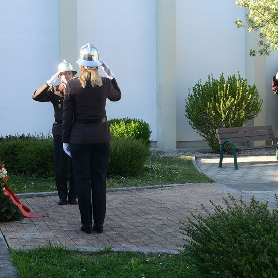 Zwei Frauen in Uniform, eine salutiert, stehen auf einem gepflasterten Bereich mit einer Bank und Grünpflanzen. Ein Gebäude mit weißen Wänden ist im Hintergrund.