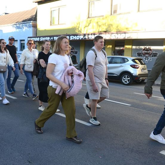 Eine Gruppe von Menschen geht vor einem Geschäft über die Straße. Einige tragen eine Brille und Sneakers, einer trägt eine rosa Jacke. Ein Auto ist in der Nähe geparkt.