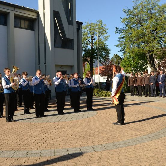 Eine Band spielt im Freien vor einem Gebäude, mit einer Person rechts stehend. Hinter der Band stehen mehrere Personen in einer Reihe. Bäume sind um den Bereich herum gepflanzt.