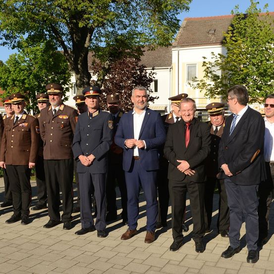 Eine Gruppe von Männern in militärischer und ziviler Kleidung steht auf einem Ziegelweg, mit Bäumen und Gebäuden im Hintergrund.
