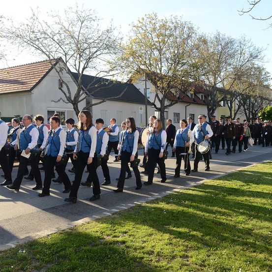 Eine Musikkapelle in blauen Westen und schwarzen Hosen marschiert eine Straße hinunter, gesäumt von Gebäuden und Bäumen.