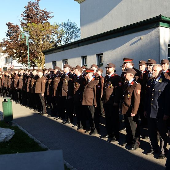 Eine Reihe von uniformierten Männern, wahrscheinlich Militär, steht in Formation auf einem Bürgersteig. Sie sind in Reihen aufgestellt und tragen Hüte und Schuhe. Dahinter ist ein weißes Gebäude mit grüner Einfassung zu sehen.