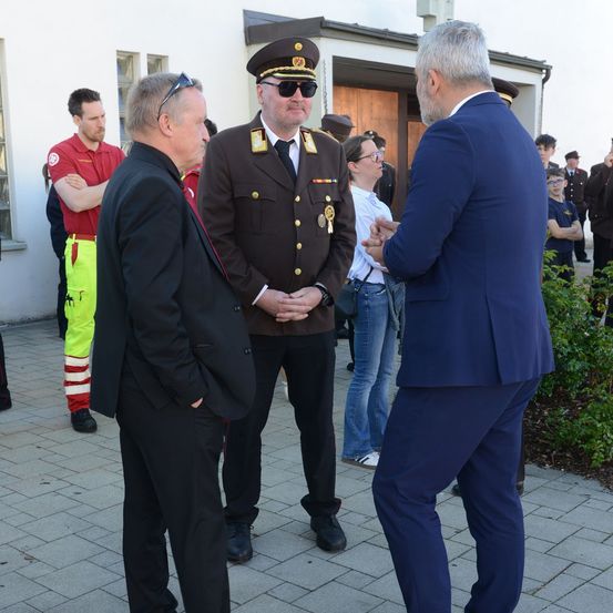 Ein Mann in Uniform steht neben einem Mann im Anzug, während ein Mann in einem roten T-Shirt zuschaut. Eine Frau steht hinter ihnen und beobachtet.