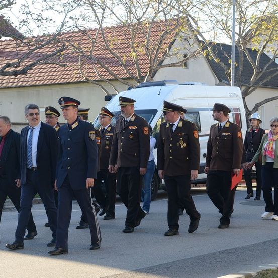 Uniformierte Offiziere und eine Gruppe Zivilisten gehen auf einer Straße, im Hintergrund sind Gebäude zu sehen. Ein Lieferwagen ist hinter ihnen geparkt.
