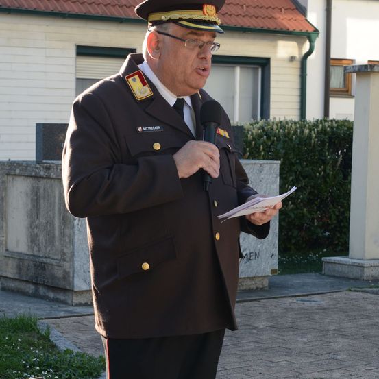 Ein Mann in Uniform hält ein Mikrofon und liest vor einem Gebäude von einem Zettel vor.