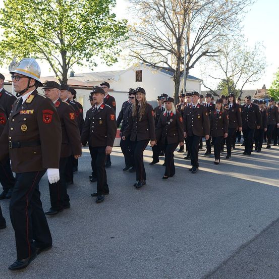 Eine Gruppe uniformierter Personen, einige mit Mützen, marschiert in einer Parade auf einer asphaltierten Straße. Bäume und Gebäude sind im Hintergrund sichtbar.