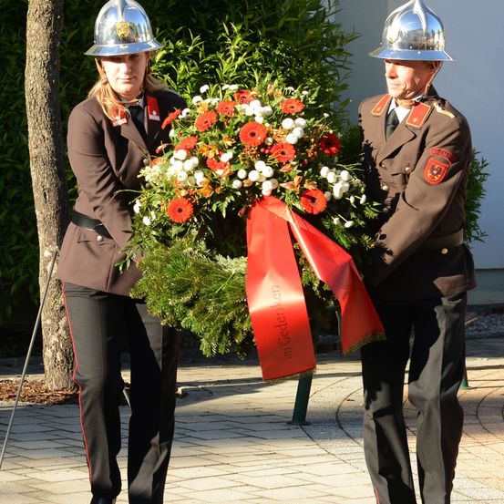 Zwei uniformierte Personen halten eine Kranz mit roten Bändern, stehen im Freien. Die Frau schaut den Mann an, beide tragen Helme. Hinter ihnen sind Bäume und ein Gebäude.