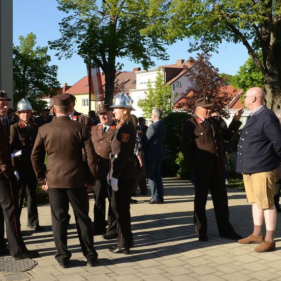Uniformierte Personen stehen in einer quadratischen Formation, einige tragen Helme und andere sind in Geschäftskleidung. Ein Mann in einem blauen Anzug gestikuliert mit seiner Hand. Gebäude sind im Hintergrund.