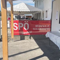 Eine Outdoor-Veranstaltung mit einem weißen Zelt. Ein roter Banner sagt 'SPO Neufeld Herzlich Willkommen'. Ein Tisch mit einem weißen Tuch. Ein Gebäude im Hintergrund.