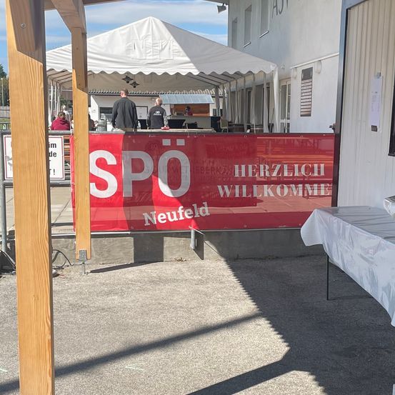 Eine Outdoor-Veranstaltung mit einem weißen Zelt. Ein roter Banner sagt 'SPO Neufeld Herzlich Willkommen'. Ein Tisch mit einem weißen Tuch. Ein Gebäude im Hintergrund.