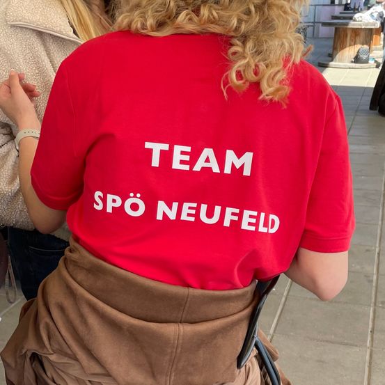 Eine Frau mit lockigem Haar trägt ein rotes T-Shirt mit weißer Schrift, die 'TEAM SPO NEUFELD' sagt.