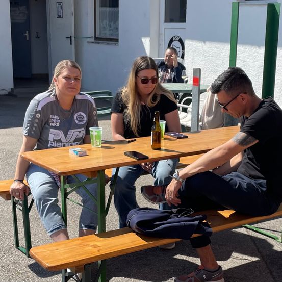 Drei Personen sitzen auf Bänken um einen Holztisch mit einer Flasche, einem Becher und einem Handy. Ein Mann in einem schwarzen T-Shirt sitzt rechts und eine Frau in einem grauen T-Shirt sitzt links. Eine weitere Frau sitzt in der Mitte.
