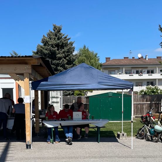 Eine Gruppe von Menschen sitzt unter einem blauen Zelt bei einer Outdoor-Veranstaltung, mit geparkten Fahrrädern in der Nähe.