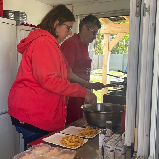 Zwei Personen kochen in einem Imbisswagen. Eine Frau in einer roten Kapuzenjacke kocht, während ein Mann in einem roten Hemd einen Kochlöffel hält. Sie sind von Tabletts mit Essen und Kochutensilien umgeben.