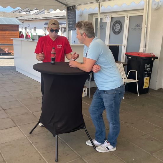 Zwei Männer stehen an einem Tisch, einer in einem roten Shirt und der andere in einem blauen Shirt. Der Mann im roten Shirt hält eine Flasche. Sie führen ein Gespräch. Dahinter steht ein Gebäude mit einem Schild.