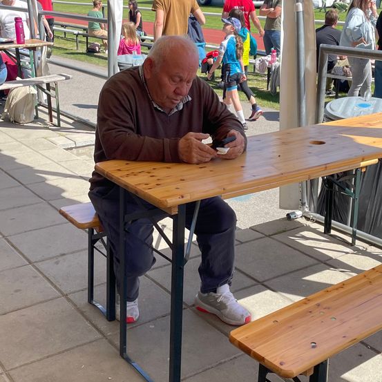 Ein älterer Mann sitzt auf einer Bank draußen, hält ein Handy in der Hand und schaut darauf. Der Tisch hat ein Loch in der Mitte. In der Ferne gehen Leute.
