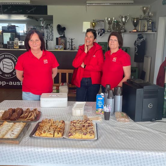 Drei Frauen in roten Shirts stehen hinter einem Tisch mit Gebäck. Eine Milchpackung und Tassen sind in der Nähe.