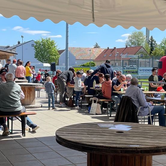 Ein überfüllter Outdoor-Essbereich mit Menschen an Tischen unter einem Zelt. Verschiedene Häuser und ein Lkw sind im Hintergrund zu sehen.