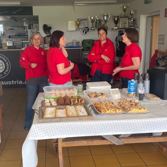 Vier Frauen in roten Jacken stehen um einen Tisch mit verschiedenen Desserts, einschließlich Gebäck, in einem Raum mit gefliesten Böden und Trophäen an der Wand.