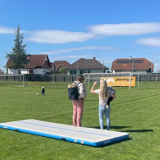 Zwei Frauen stehen neben einem Fußballfeld mit einer blau-grauen aufblasbaren Matte. Ein Fußballer in der Ferne tritt einen Ball. Häuser mit Solarpanelen und ein Baum sind im Hintergrund zu sehen.