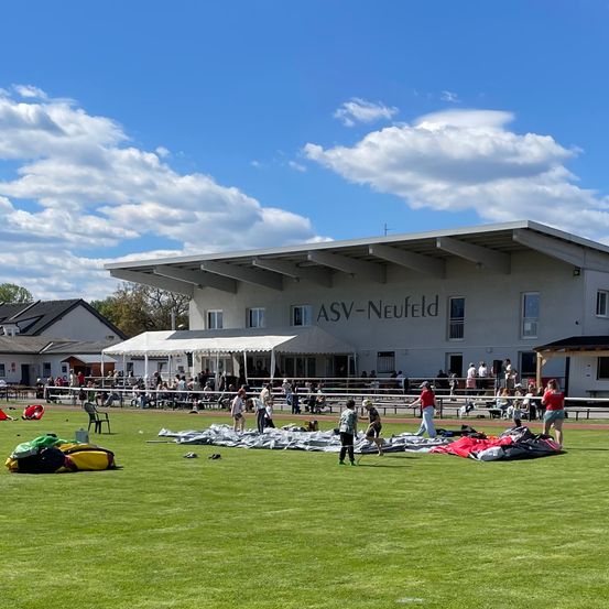 Ein Outdoor-Event am ASV-Neufeld-Gelände mit einer Gruppe von Menschen bei verschiedenen Aktivitäten, einem großen weißen Gebäude und einem Vordach. Menschen bauen Zelte und Ausrüstung auf dem Rasenfeld auf.