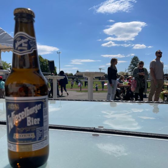Eine Flasche Wiesellburger Bier steht auf einem Tisch in einer Außenumgebung, mit Menschen im Hintergrund unter einem teilweise bewölkten Himmel.
