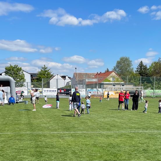 Eine Gruppe von Menschen spielt auf einem großen grünen Feld. Einige spielen Fußball, während andere verschiedene Aktivitäten ausführen. Ein paar Häuser und Bäume sind im Hintergrund sichtbar.