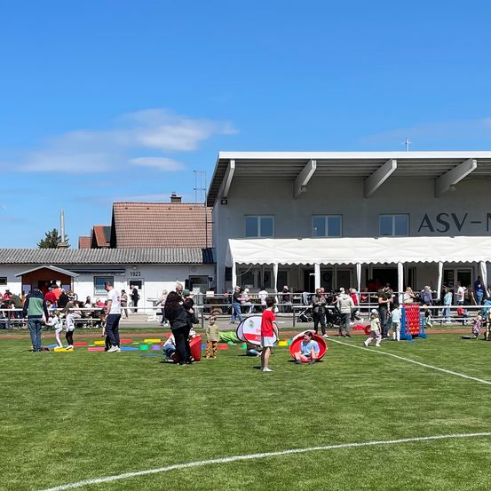 Viele Menschen genießen Outdoor-Aktivitäten auf einem Sportplatz, mit einem großen Gebäude mit der Aufschrift ASV-N im Hintergrund.