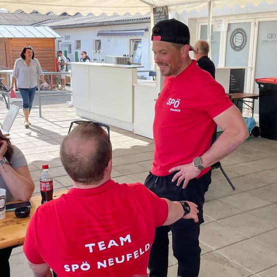 Zwei Männer in roten Team Spö Neufeld-Shirts stehen in einer öffentlichen Umgebung, wahrscheinlich an einem Imbissstand. Der Mann auf der linken Seite hat den Rücken zur Kamera, und der andere Mann lächelt. In der Nähe laufen Menschen, und ein Gebäude ist zu sehen.