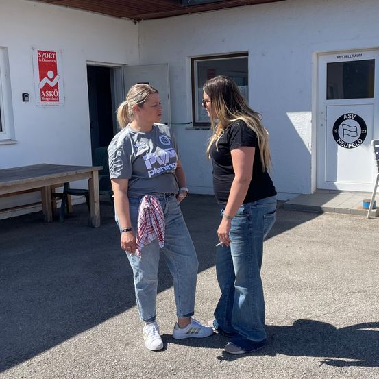 Zwei Frauen stehen vor einem Gebäude mit einem Schild, das Sport in Österreich anzeigt. Eine Frau raucht eine Zigarette und die andere trägt eine Uhr.
