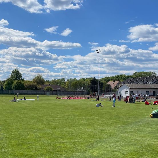 Ein großes grünes Feld mit mehreren Personen, die verschiedene Aktivitäten ausführen, einem Gebäude im Hintergrund und einem strahlend blauen Himmel mit Wolken.
