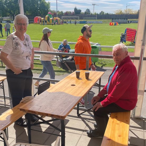 Eine Gruppe von Menschen sitzt an einem Holztisch in einem überdachten Bereich, mit einem Mann in einem roten Pullover, der allein auf einer Bank sitzt. Eine Frau mit Brille und ein Mann in einem orangefarbenen Hoodie schauen sich gegenseitig an. Eine Frau geht im Hintergrund.