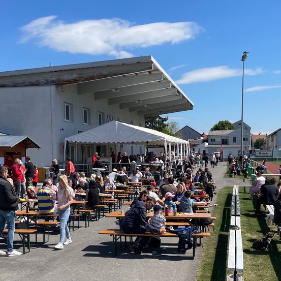 Freisitz bei einer Sportanlage mit vielen versammelten Menschen, einige unter einem großen Zelt. Ein weißes Gebäude mit dem Namen 'ASV' im Hintergrund.
