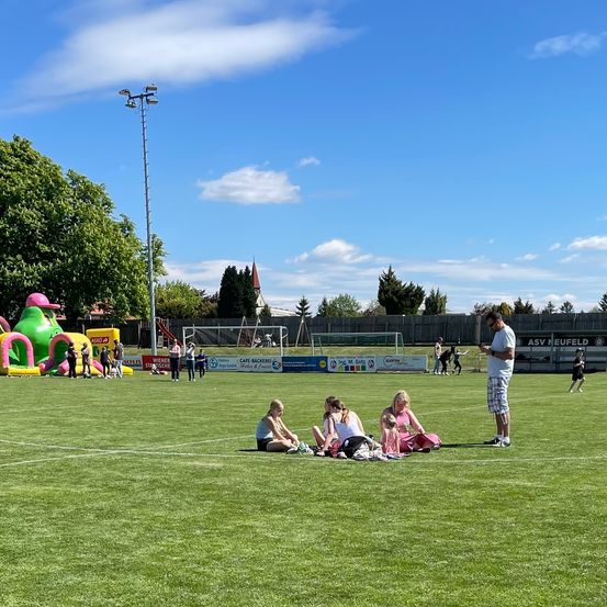 Ein Fußballplatz mit Familien auf dem Rasen, einer Person, die ihr Telefon benutzt, und einem aufblasbaren Spielplatz in der Ferne.