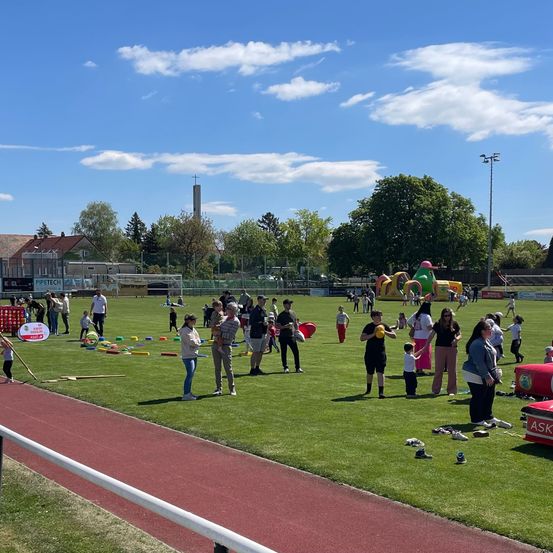 Ein sonniger Tag bei einer Veranstaltung im Freien, mit Familien, die auf einem Feld Sport treiben und aufblasbare Spiele im Hintergrund. Bäume und Gebäude sind in der Ferne sichtbar.