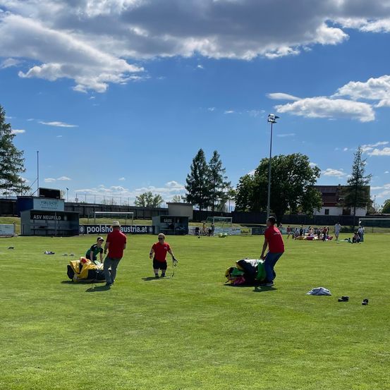 Ein sonniger Tag mit Menschen auf einem Feld. Einige richten Ausrüstung auf, andere versammeln sich in Gruppen. Bäume und ein Gebäude sind im Hintergrund.