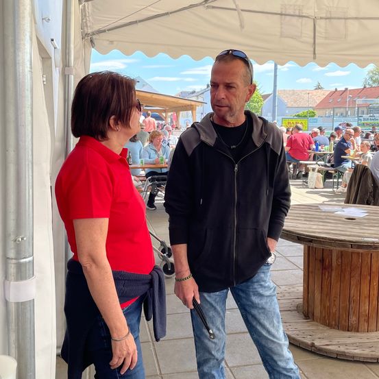 Ein Mann in einer schwarzen Jacke und Sonnenbrille hält einen Stock und steht neben einer Frau in einem roten Shirt, unter einem weißen Baldachin. Ein hölzerner Tisch ist in der Nähe.