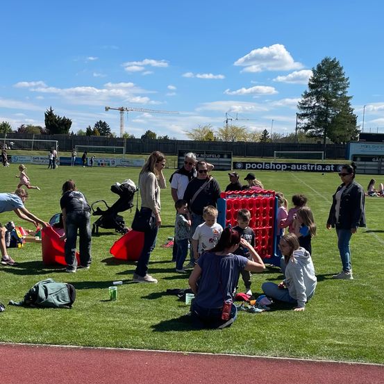 An einem sonnigen Tag spielt eine Gruppe von Familien auf einem Sportplatz. Einige Kinder spielen ein Spiel, während andere zuschauen. Einige Erwachsene stehen und reden.