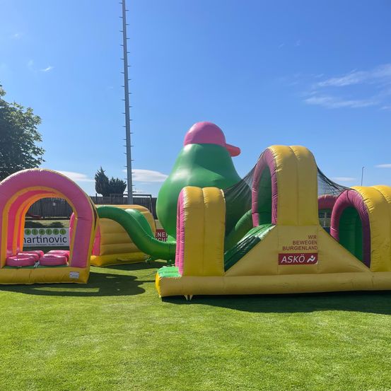 Ein aufblasbarer Spielplatz mit einem großen Vogel und verschiedenen Hindernissen steht auf einem grünen Feld unter klarem Himmel.