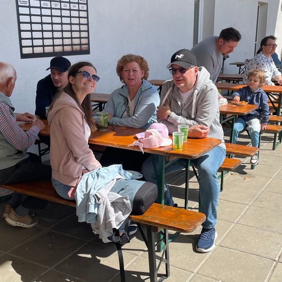 Eine Gruppe von Menschen sitzt an Tischen im Freien und lächelt. Zwei Frauen tragen Sonnenbrillen, eine in einer rosa Jacke. Ein Kind sitzt in einer blauen Jacke und hält etwas zu essen. Ein Mann trägt eine graue Kapuzenjacke und Sonnenbrille. Die Tische haben Gläser und Jacken.