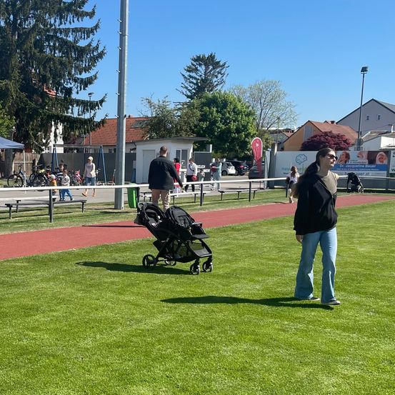 Ein Rasenfeld mit einer Laufbahn. Eine Frau mit einem Kinderwagen und ein Mann gehen an einer Bank vorbei. Bäume, ein Gebäude und ein Auto sind im Hintergrund.