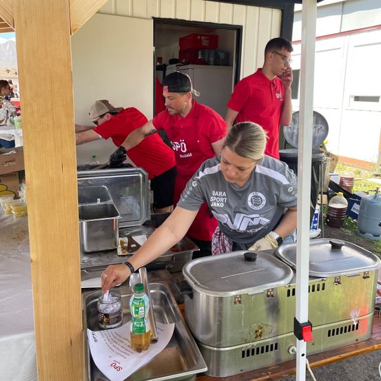 Eine Frau in einem grauen Shirt bereitet Essen an einem Imbissstand vor, umgeben von mehreren Männern in roten Shirts. Sie hält eine Flasche und Papier, mit zwei großen Kochtöpfen auf dem Stand.