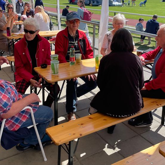 Eine Gruppe älterer Menschen sitzt an einem Holztisch bei einer Veranstaltung im Freien. Sie sind in Freizeitkleidung gekleidet und tragen Sonnenbrillen. Auf dem Tisch stehen Becher und eine Flasche.