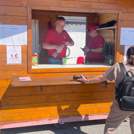 Zwei Männer in roten Shirts arbeiten in einem kleinen Kiosk. Einer hält eine Flasche Wasser, der andere schaut auf ein Handy. Eine Kundin überreicht Bargeld. Ein Menübrett mit Getränken und Preisen ist sichtbar.