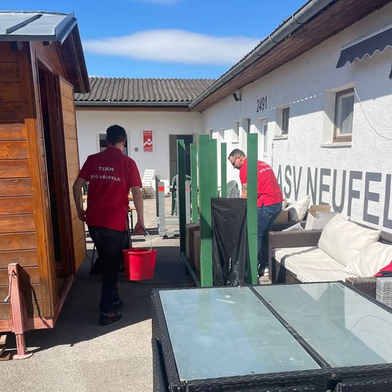 Zwei Männer in roten Uniformen reinigen vor einem Gebäude mit der Aufschrift ASV Neufeld. Einer hält einen roten Eimer, während der andere sich über einen grünen Zaun beugt. Ein Sofa und ein Tisch mit Glasplatte befinden sich in der Nähe.