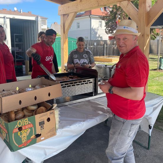 Eine Gruppe von Menschen in roten Shirts grillt Essen unter einem hölzernen Pavillon. Eine Frau in Grau bereitet Essen zu, während ein Mann mit Hut zuschaut.