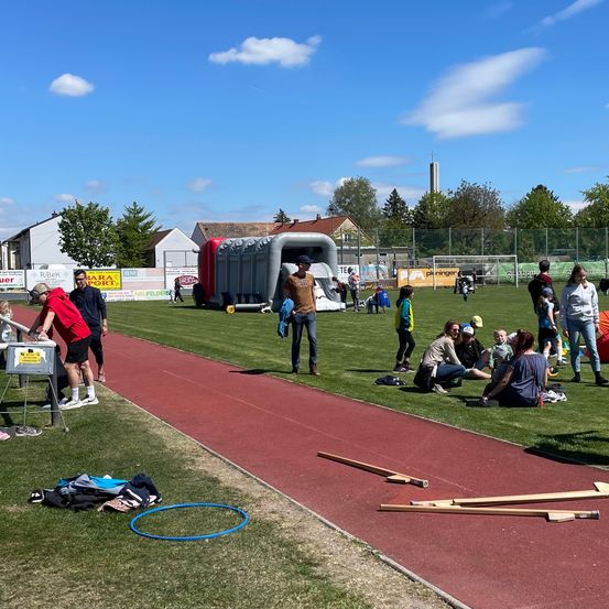 Eine Leichtathletikveranstaltung ist im Gange, mit Teilnehmern in verschiedenen Vorbereitungsphasen. Mehrere Personen sitzen an der Seite der Bahn, während andere stehen. Ausrüstung und Kleidung sind verstreut.