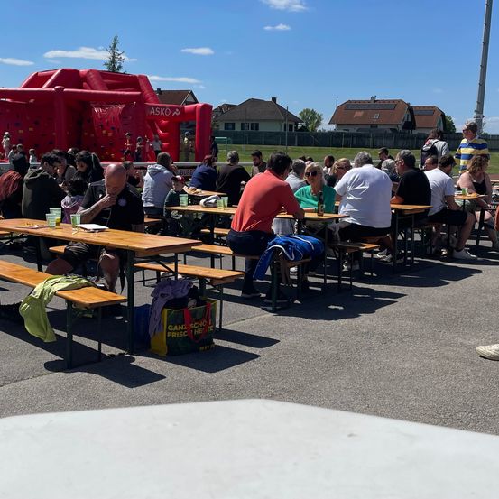 Eine Gruppe von Menschen sitzt an Picknicktischen im Freien unter einem blauen Himmel, in der Nähe einer aufblasbaren Spielstruktur. Sie haben Gläser und Essen auf den Tischen.