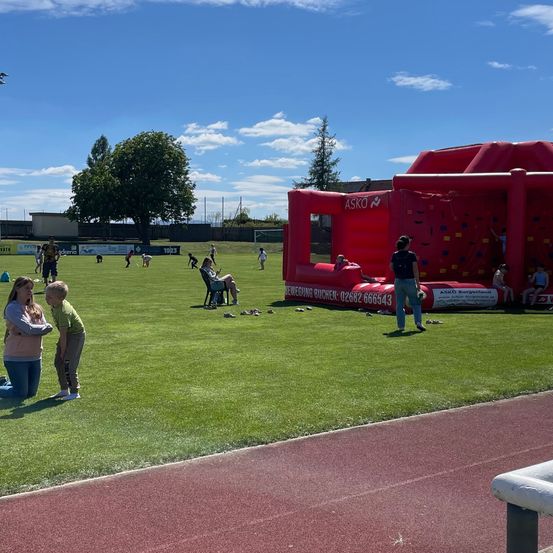 Ein Freiluftsportplatz mit grünem Rasen, einer roten aufblasbaren Struktur und spielenden und herumstehenden Menschen. Bäume und ein Zaun sind im Hintergrund sichtbar.